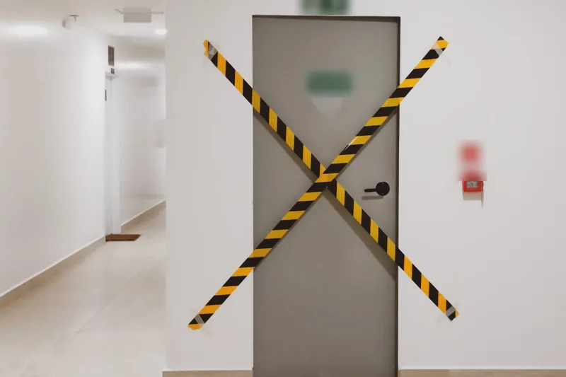 Closed door in a bright hallway crossed with yellow and black hazard tape (hazard sign text blurred)